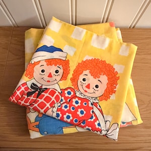 Puede incluir: Sábanas vintage amarillas con un patrón de cuadrados blancos repetidos. Las sábanas están decoradas con un diseño de muñecas Raggedy Ann y Andy. Las muñecas tienen pelo rojo de hilo, mejillas rosadas y visten trajes coloridos.