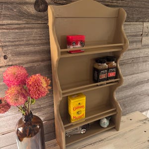May include: A beige-painted wooden spice rack with four shelves. The rack holds various spice containers, including a red tin, yellow mustard tin, and two Trader Joe's spice jars. A small ceramic bird and measuring spoons are also visible.