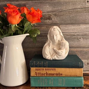Könnte beinhalten: Eine weiße Keramikfigur der Jungfrau Maria, die ein Jesuskind hält. Die Figur steht auf einem Stapel von drei Büchern. Das oberste Buch trägt den Titel "Attachments" von Judith Rossner. Das mittlere Buch trägt den Titel "The In Epistle of Saint Paul to the Corinthians" von Jean Hering. Das unterste Buch trägt den Titel "The Chinese Looking Glass" von Dennis Bloodworth. Die Bücher stehen auf einer Holzoberfläche.