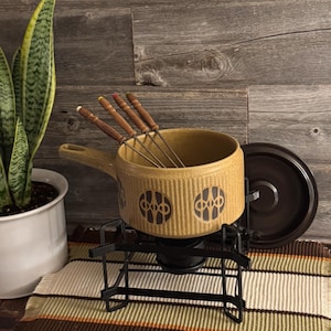 Puede incluir: Un juego de fondue vintage con una olla de cerámica beige con un diseño geométrico negro. El juego incluye un soporte de metal negro, una tapa a juego y tenedores de fondue con mangos de madera.