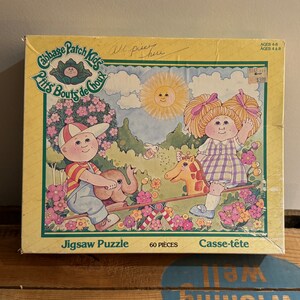 May include: A 60-piece jigsaw puzzle featuring two Cabbage Patch Kids dolls on a seesaw. The girl doll is wearing a purple and white checked dress and has blonde pigtails. The boy doll is wearing a red and white striped shirt and a blue baseball cap. The puzzle box has a yellow background with green borders and the text "Cabbage Patch Kids" and "Jigsaw Puzzle" in large, bold letters.