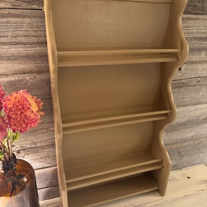 May include: A light beige wooden wall shelf with four horizontal shelves. The shelf has a curved, decorative edge and is painted in a neutral tone. The background features a wooden wall and a vase of flowers.