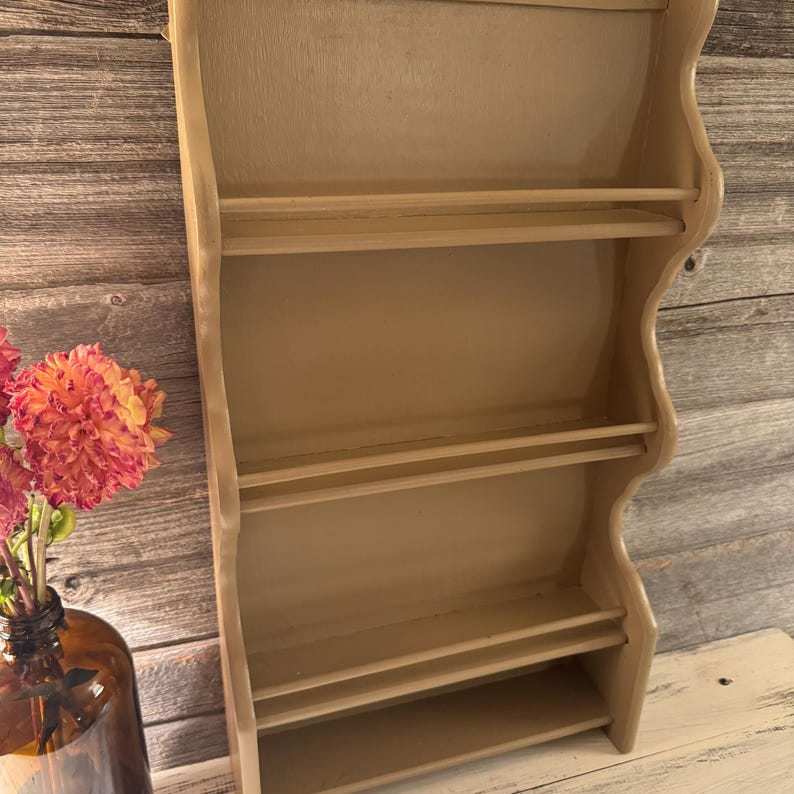 May include: A light beige wooden wall shelf with four horizontal shelves. The shelf has a curved, decorative edge and is painted in a neutral tone. The background features a wooden wall and a vase of flowers.