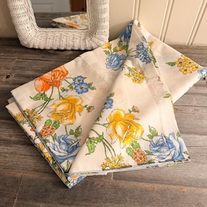 May include: Cream-coloured floral fabric featuring large yellow, orange, and blue roses. The fabric is folded, displaying the floral pattern. The flowers are surrounded by green leaves and smaller yellow and blue blossoms. The fabric is likely a tablecloth or bedding.