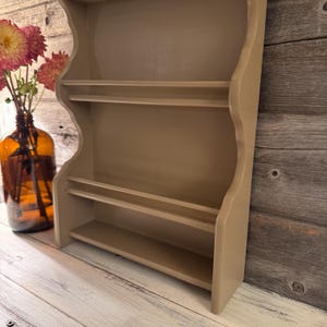May include: A beige-colored, wall-mounted shelf unit with three shelves. The unit has a scalloped edge design and is made of wood. A brown glass bottle with flowers is to the left of the shelf.