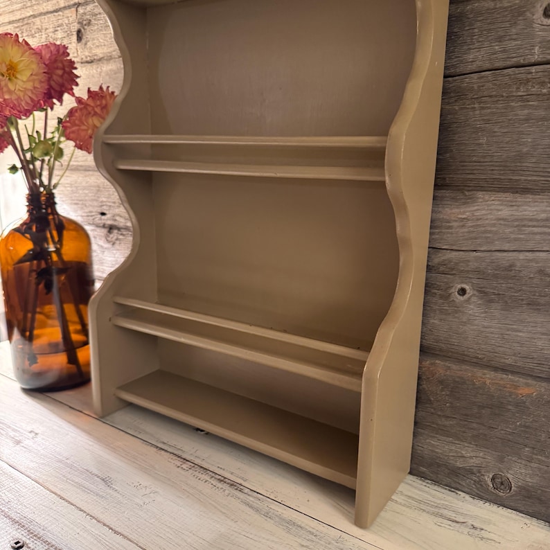 May include: A beige-colored, wall-mounted shelf unit with three shelves. The unit has a scalloped edge design and is made of wood. A brown glass bottle with flowers is to the left of the shelf.