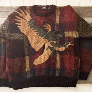 Peut inclure: Un pull en laine à carreaux marrons, noirs et beiges avec une applique d'aigle marron. Le pull a un col rond et des manches longues.