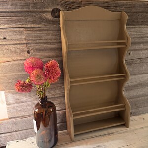 May include: A beige wooden wall shelf with three shelves and a curved top. A brown glass bottle with a bouquet of pink and orange flowers sits to the left of the shelf. The background is a rustic wooden wall.