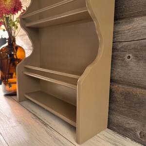 May include: A beige-colored, wooden wall shelf with three shelves. The shelf has a decorative, scalloped edge and is designed for displaying small items. The shelf is painted in a neutral tone, suitable for various decor styles. The shelf is shown from a side angle.