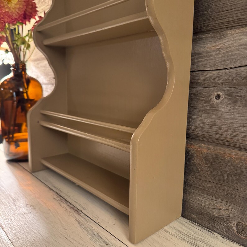 May include: A beige-colored, wooden wall shelf with three shelves. The shelf has a decorative, scalloped edge and is designed for displaying small items. The shelf is painted in a neutral tone, suitable for various decor styles. The shelf is shown from a side angle.