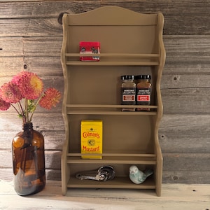 May include: A beige wooden spice rack with four shelves, holding various spice containers, including a red tin, yellow Colman's Mustard tin, and Trader Joe's spice jars. A small bird figurine and measuring spoons are on the bottom shelf.