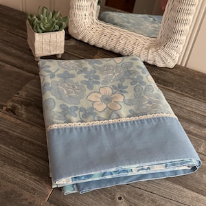 Op de afbeelding: Een blauw en wit beddengoed met een bloemenpatroon en een witte kantversiering. Het beddengoed is gevouwen en ligt op een houten oppervlak.