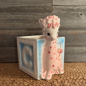 Könnte beinhalten: Keramik-Babyblock-Pflanzgefäß mit Giraffen-Design. Der Block ist weiß mit hellblauem Rand und dem Buchstaben "C". Die Giraffe ist rosa mit roten Tupfen und einem rosa Kopf.