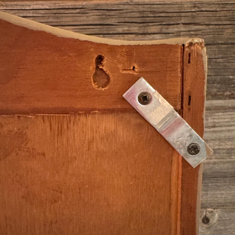 May include: Close-up of a wooden frame corner with a metal bracket attached. The wood is a warm brown color, and the bracket is secured with two screws. A keyhole is visible.