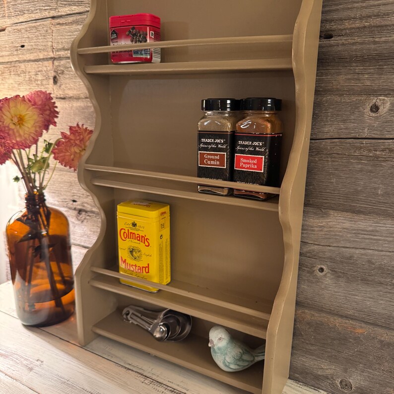 May include: A beige, wall-mounted spice rack with multiple shelves. It holds various items, including spice jars, a yellow Coleman's Mustard tin, and a small bird figurine. A vase of pink flowers sits to the left. The rack is against a wooden background.