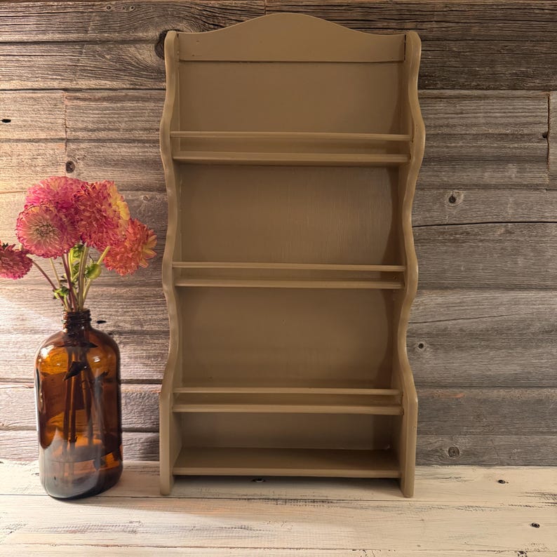 May include: A beige-colored, wooden spice rack with four shelves. The rack has a decorative, curved top and sides. A brown glass bottle with pink and orange flowers sits to the left of the rack, adding a touch of color to the rustic scene.