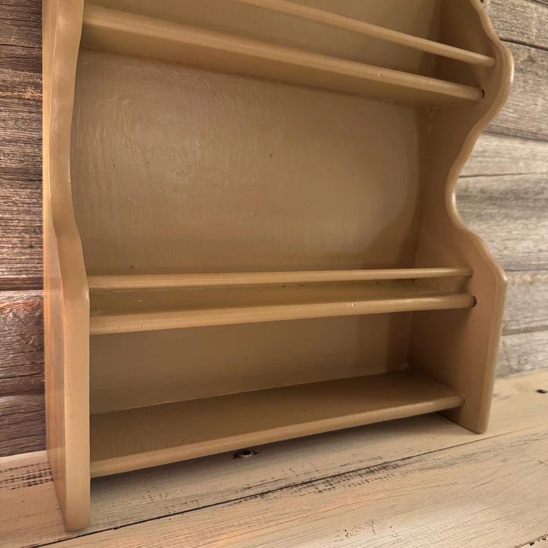 May include: A light beige wooden spice rack with three shelves and a curved design. The rack is mounted on a light-colored wooden surface, with a weathered wood background.