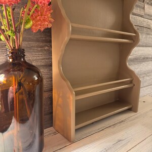 May include: A beige-painted wooden wall shelf with three shelves and a decorative wavy edge. The shelf is empty and stands on a white surface. A brown glass bottle with orange flowers is to the left of the shelf.