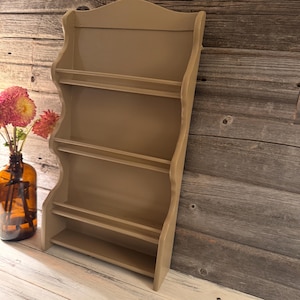 May include: A beige-colored, wooden wall shelf with a scalloped edge design. The shelf has four tiers for displaying items. A brown glass bottle with pink flowers is to the left of the shelf.