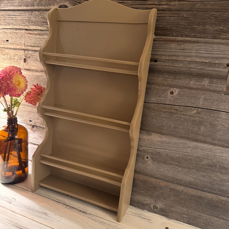 May include: A beige-colored, wooden wall shelf with a scalloped edge design. The shelf has four tiers for displaying items. A brown glass bottle with pink flowers is to the left of the shelf.