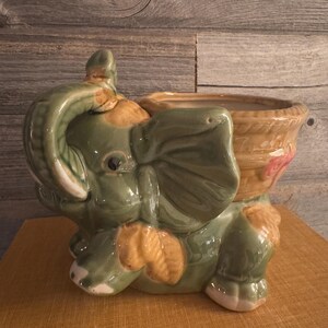 Puede incluir: Una maceta de cerámica con forma de elefante en tonos verdes, beige y crema. El elefante está sentado con la trompa enrollada. La maceta tiene un recipiente en forma de cesta en la espalda, con un diseño floral rosa.