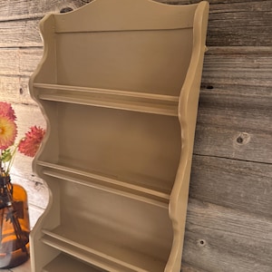 May include: A beige-colored, wall-mounted spice rack with four shelves. The rack has a decorative, scalloped top and sides. The shelves are designed to hold small jars or containers. The rack is made of wood and has a painted finish.