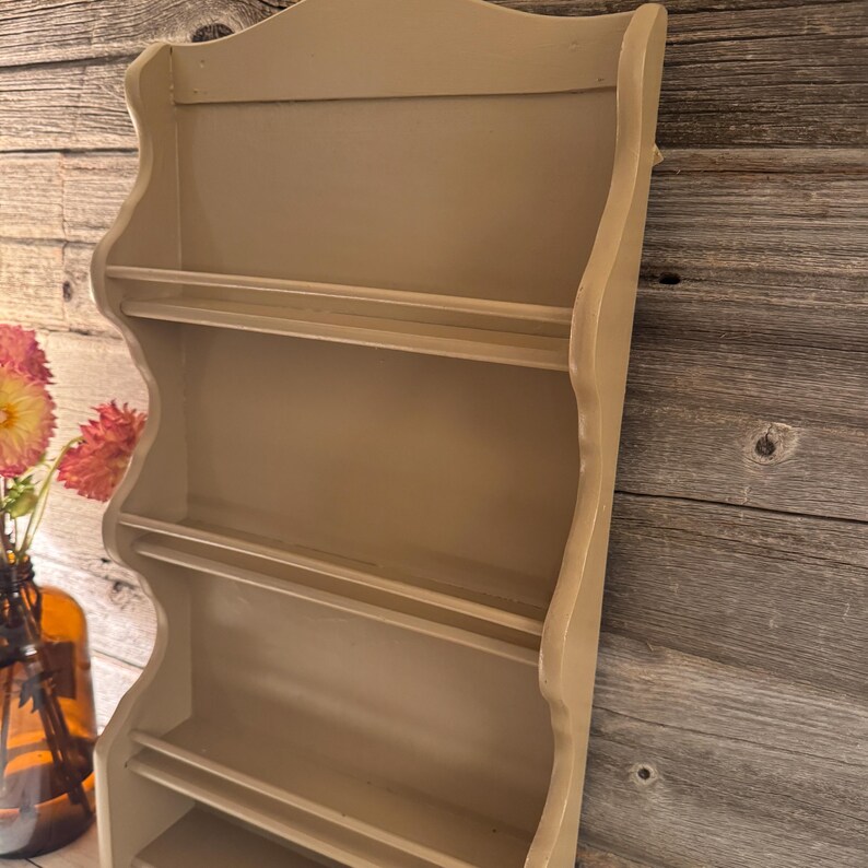 May include: A beige-colored, wall-mounted spice rack with four shelves. The rack has a decorative, scalloped top and sides. The shelves are designed to hold small jars or containers. The rack is made of wood and has a painted finish.