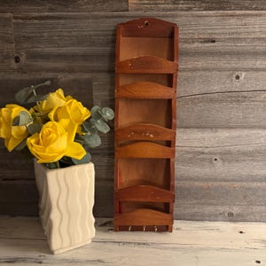 Peut inclure: Un organiseur de courrier en bois vintage avec cinq fentes et trois petits crochets en bas. L'organisateur est de couleur marron chaud et a une finition vieillie. Un vase blanc avec des roses jaunes et de l'eucalyptus est à gauche.