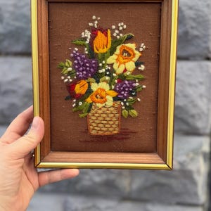 Può includere: Opera d'arte floreale ricamata in una cornice di legno. L'opera presenta un bouquet di fiori colorati, tra cui tulipani arancioni, narcisi gialli e uva viola, disposti in un cestino intrecciato. La cornice ha un bordo interno dorato.