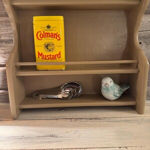 May include: A beige wooden spice rack with two shelves. The top shelf holds a yellow tin of Colman's Mustard. The bottom shelf has a set of measuring spoons and a small blue bird figurine.