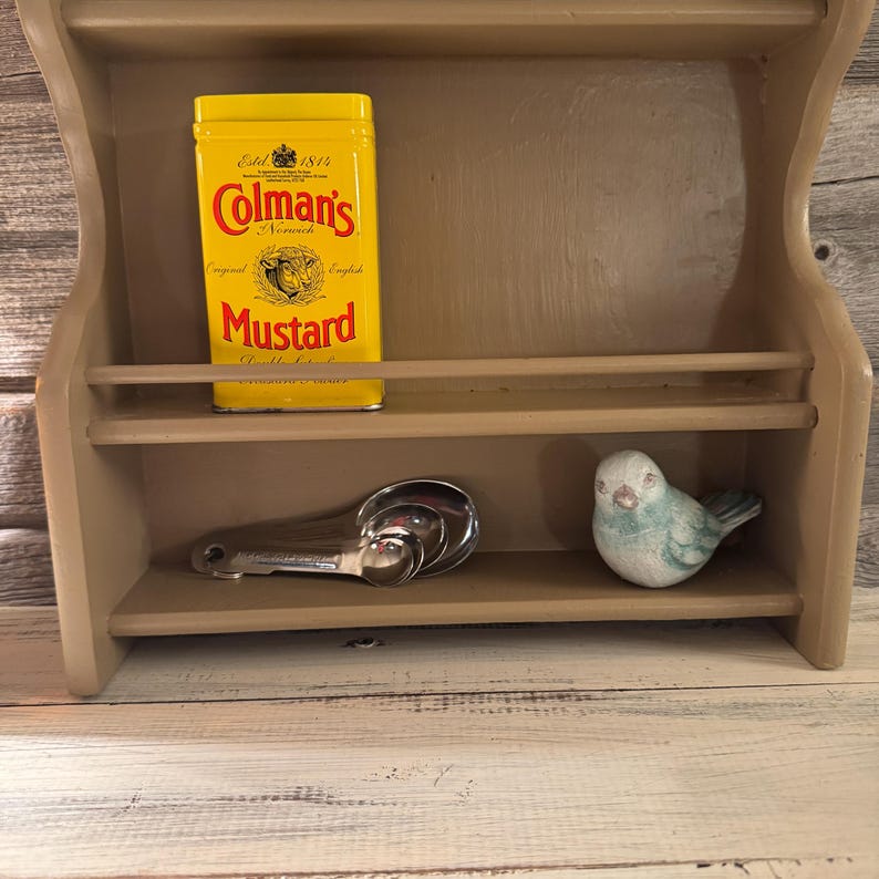 May include: A beige wooden spice rack with two shelves. The top shelf holds a yellow tin of Colman's Mustard. The bottom shelf has a set of measuring spoons and a small blue bird figurine.