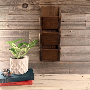 May include: A dark brown wooden wall-mounted organiser featuring three vertical storage pockets with a curved side profile. The unit has a smooth, stained finish that highlights the natural wood grain texture throughout the entire structure.
