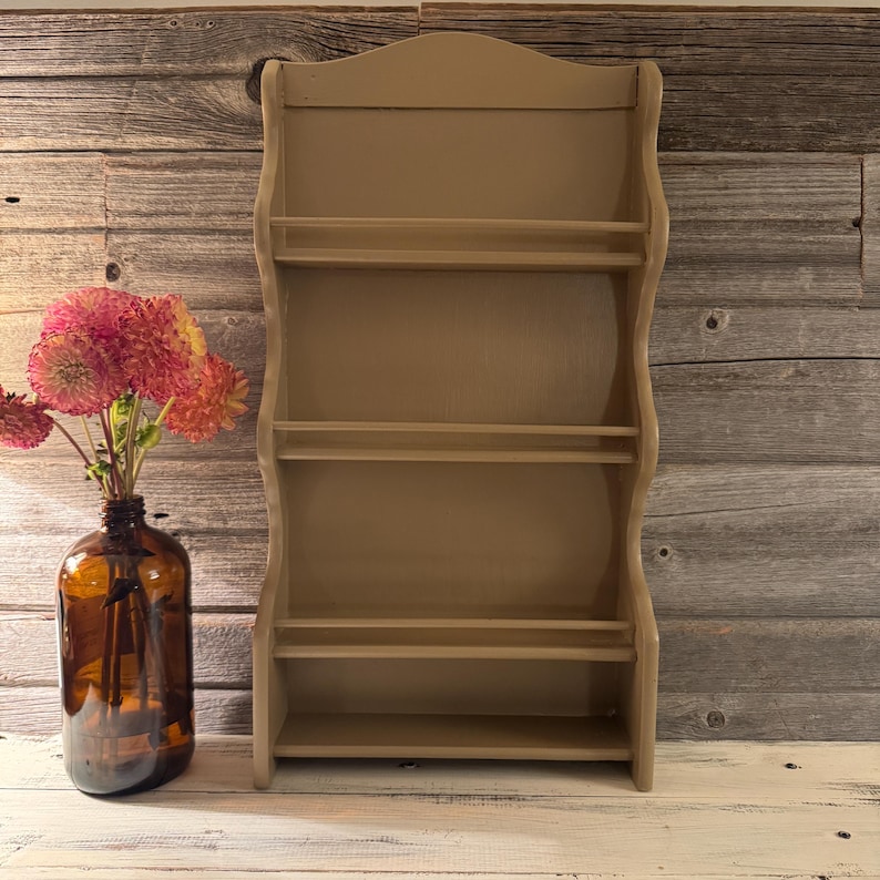 May include: A beige-colored, wall-mounted spice rack with four shelves. The rack has a curved design and is made of wood. A brown glass bottle with pink and orange flowers is to the left of the rack.