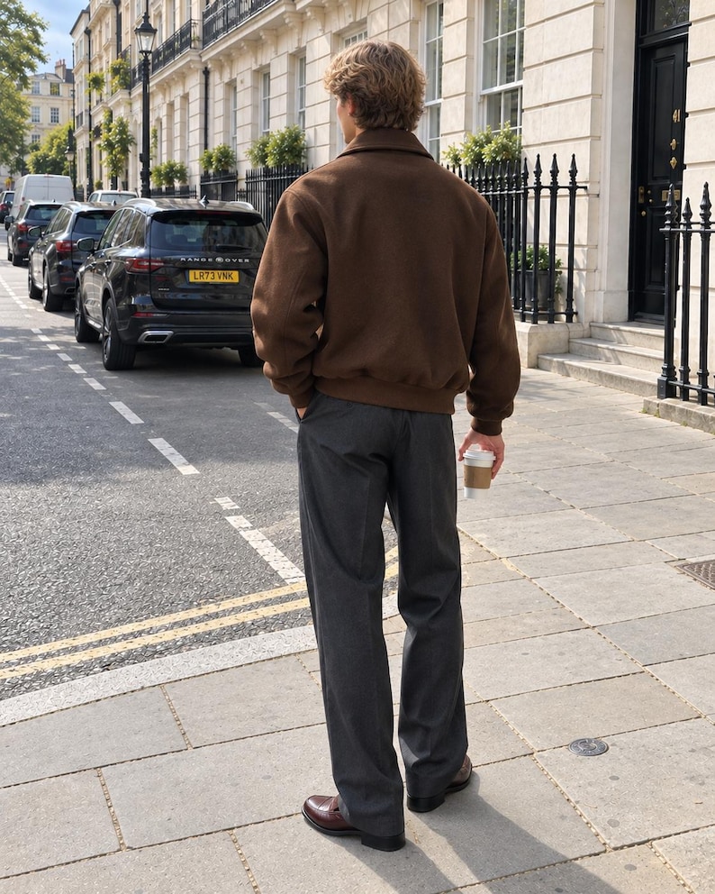 May include: A brown collared jacket with long sleeves and ribbed cuffs, paired with charcoal gray trousers and dark brown leather loafers. The outfit is worn with a white paper coffee cup held in one hand, showcasing a clean and structured look.
