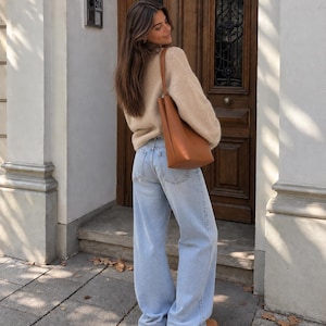 May include: A beige long-sleeved knit sweater, light blue denim jeans with a wide-leg cut, a tan leather shoulder bag, and tan platform shoes. The outfit is worn together, showcasing the texture of the knit fabric and the light wash of the denim.