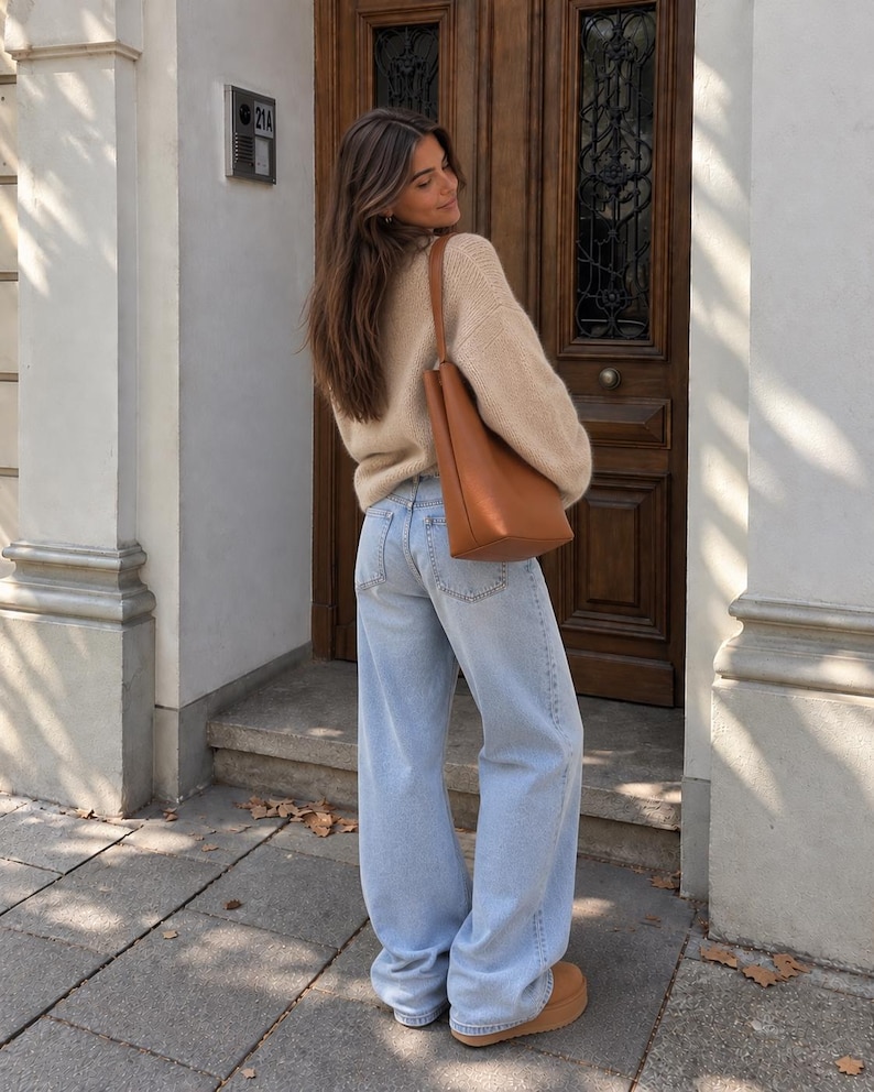 May include: A beige long-sleeved knit sweater, light blue denim jeans with a wide-leg cut, a tan leather shoulder bag, and tan platform shoes. The outfit is worn together, showcasing the texture of the knit fabric and the light wash of the denim.