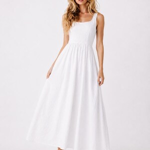 May include: A floor-length white sleeveless dress featuring a square neckline, a fitted bodice, and a full, flowing skirt. The fabric has a subtle, textured appearance, and the garment is paired with simple beige heeled sandals.