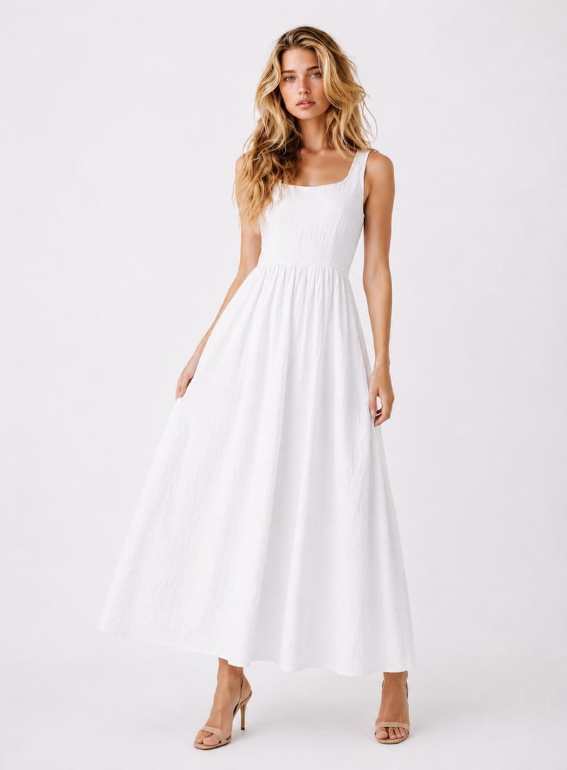 May include: A floor-length white sleeveless dress featuring a square neckline, a fitted bodice, and a full, flowing skirt. The fabric has a subtle, textured appearance, and the garment is paired with simple beige heeled sandals.