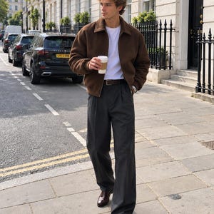 May include: A brown zip-up jacket with a collar, a plain white t-shirt, dark gray pleated trousers, a brown leather belt, and dark brown leather loafers. The outfit is worn with a silver wristwatch and a white disposable coffee cup is held.