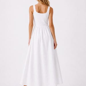 May include: A sleeveless white maxi dress featuring a square neckline and a textured fabric finish. The dress has a fitted bodice that transitions into a full, flowing skirt that reaches the ankles, paired with light tan heeled sandals.