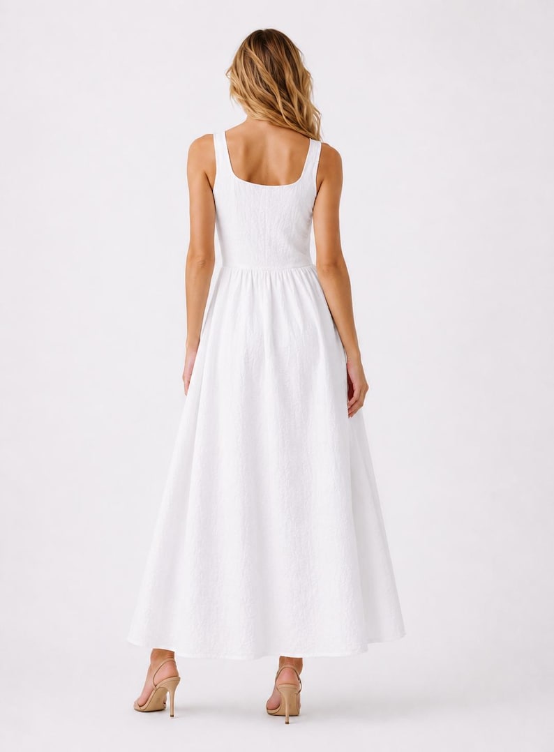 May include: A sleeveless white maxi dress featuring a square neckline and a textured fabric finish. The dress has a fitted bodice that transitions into a full, flowing skirt that reaches the ankles, paired with light tan heeled sandals.