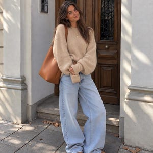 May include: A beige long-sleeved knit sweater with a crew neckline and ribbed cuffs, paired with light-wash wide-leg denim jeans, a tan leather shoulder bag, and tan suede platform shoes. The outfit is worn with a simple gold pendant necklace.
