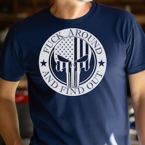 以下が含まれることがあります： ネイビーブルーのTシャツ。白いグラフィックは、アメリカ国旗を組み込んだスカルデザインで、「FUCK AROUND AND FIND OUT」の文字がスカルの周りにプリントされています。
