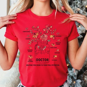 May include: A red t-shirt featuring a floral design in a jar with the word "DOCTOR" and the phrase "Helping you grow to your full potential." The design includes words like "Compassion," "Stamina," and "Patience."