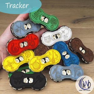 Op de afbeelding: Een verzameling houten speltrackers in verschillende kleuren, waaronder groen, blauw, rood en geel. Elke tracker heeft een uniek gegraveerd ontwerp en een draaiknop met cijfers. Het woord "Tracker" staat linksboven.