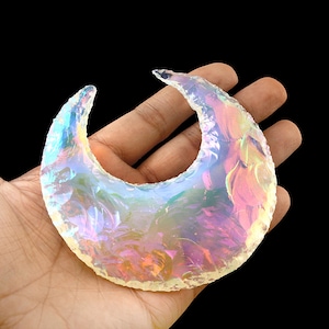 May include: A crescent-shaped, iridescent crystal. The crystal displays a range of colors, including pink, blue, and yellow, with a translucent quality. The edges are rough and uneven, giving it a natural, handcrafted appearance. The crystal is held in a hand against a black background.