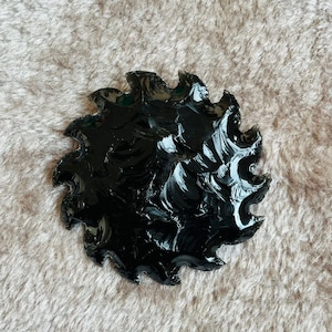 May include: A glossy, black obsidian arrowhead with a jagged, circular design. The edges are sharply defined, and the surface reflects light, showcasing the natural texture and craftsmanship of the stone.
