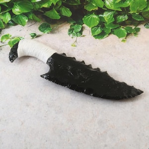 May include: A black obsidian knife with a jagged edge and a white wrapped handle. The blade is a deep, glossy black, and the handle is tightly wrapped with white cord. The knife is displayed against a neutral background with green foliage.