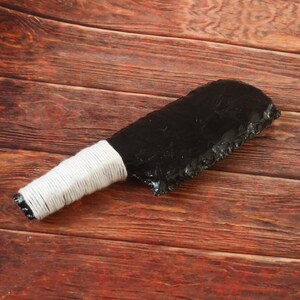 May include: A black obsidian knife with a white wrapped handle. The blade has a rough, textured edge. The handle is wrapped with white twine. The knife is displayed on a wooden surface.