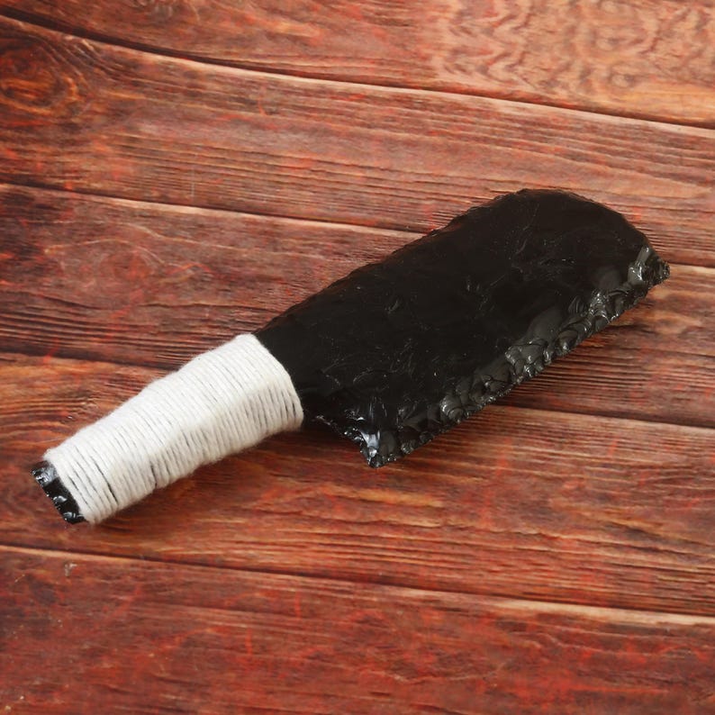May include: A black obsidian knife with a white wrapped handle. The blade has a rough, textured edge. The handle is wrapped with white twine. The knife is displayed on a wooden surface.